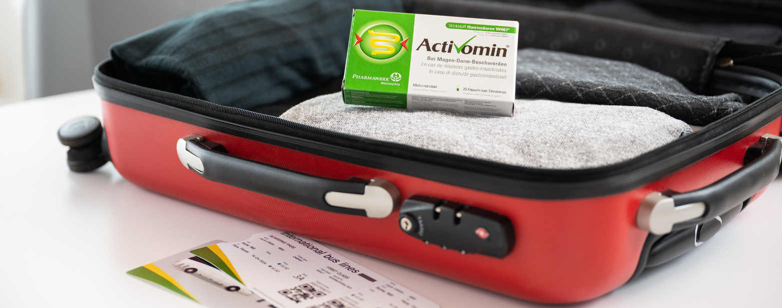 Activomin-Schachtel steht in einem geöffnetem roten Koffer auf Kleidung, davor liegt ein Bus-Ticket